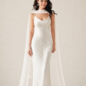 May include: A floor-length white satin slip dress featuring thin spaghetti straps and a V-neckline. The dress is adorned with delicate white floral embroidery on the side and is paired with a long, sheer, flowing white chiffon cape-style scarf.