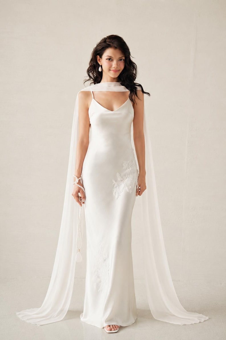 May include: A floor-length white satin slip dress featuring thin spaghetti straps and a V-neckline. The dress is adorned with delicate white floral embroidery on the side and is paired with a long, sheer, flowing white chiffon cape-style scarf.