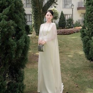 Puede incluir: Un vestido largo de color crema con una capa superpuesta transparente, escote redondo y falda fluida. La prenda se combina con un collar largo de cuentas y un pequeño bolso de mano acolchado de acabado metálico con herrajes plateados.