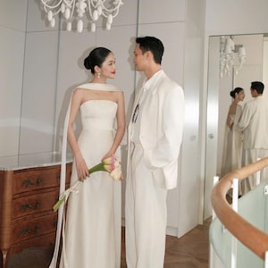 May include: A couple in formal attire, likely for a wedding. The woman wears a white strapless gown and holds flowers. The man is in a white suit. A chandelier hangs above them. The scene is reflected in a mirror.