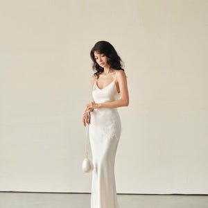 May include: A floor-length ivory satin slip dress with thin spaghetti straps, a V-neckline, and delicate white floral lace appliqu&eacute; detailing on the left hip. The dress is paired with a small, round, white beaded handbag with a pearl-like finish.