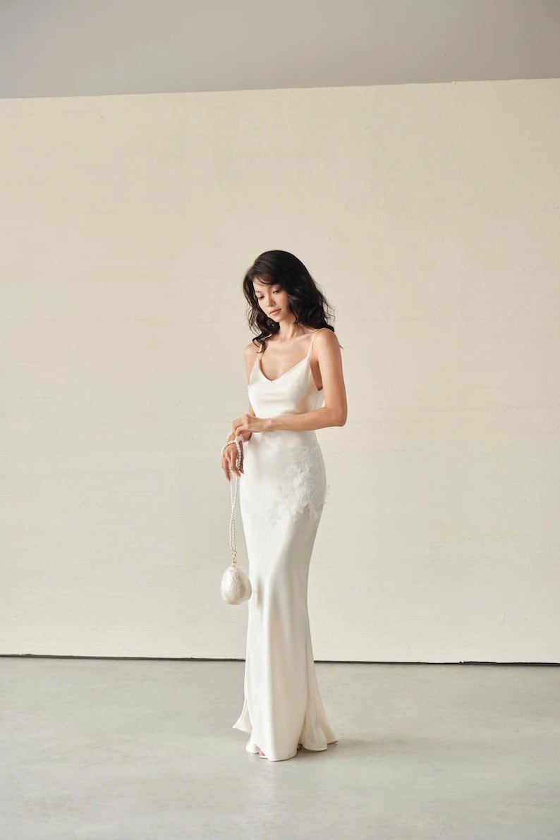 May include: A floor-length ivory satin slip dress with thin spaghetti straps, a V-neckline, and delicate white floral lace appliqu&eacute; detailing on the left hip. The dress is paired with a small, round, white beaded handbag with a pearl-like finish.