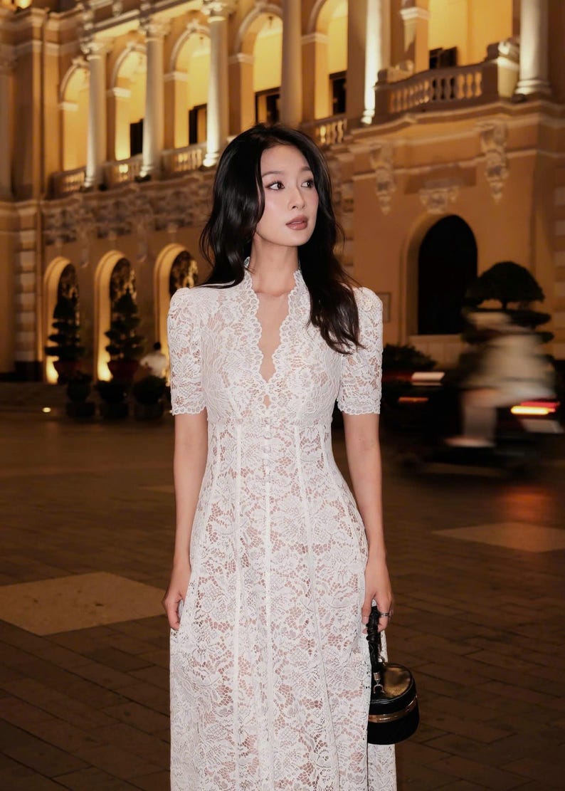 May include: A white lace dress with short sleeves and a V-neck. The dress is knee-length and has a fitted silhouette. The background features a building with lights.