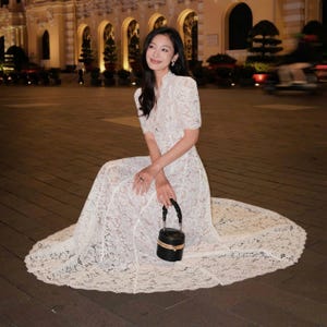May include: A woman wearing a white lace dress with short sleeves and a high neckline, sitting on a paved surface. She holds a small, black, round handbag with a gold trim. The dress has a flowing skirt.