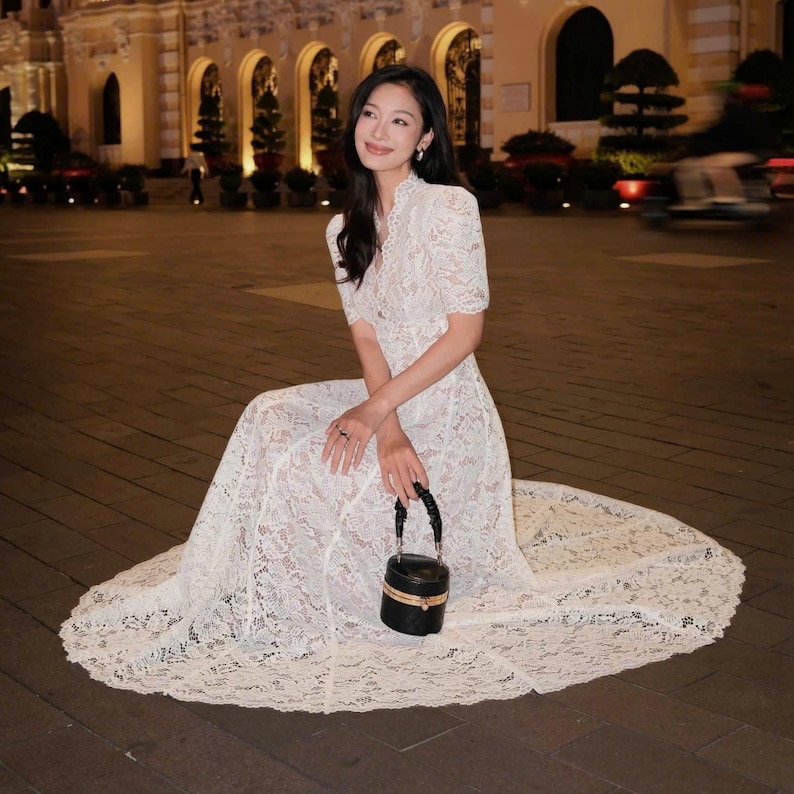 May include: A woman in a white lace dress with short sleeves and a high neckline, seated on a paved surface. She holds a small, black, round handbag with a gold trim. The dress features a flowing skirt.