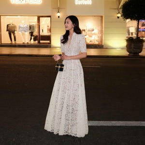 May include: A woman in a white lace dress with short sleeves and a V-neck. The dress has a long, flowing skirt. She is carrying a small, black and gold handbag. The background shows a shopfront with the Tom Ford and Berluti logos.