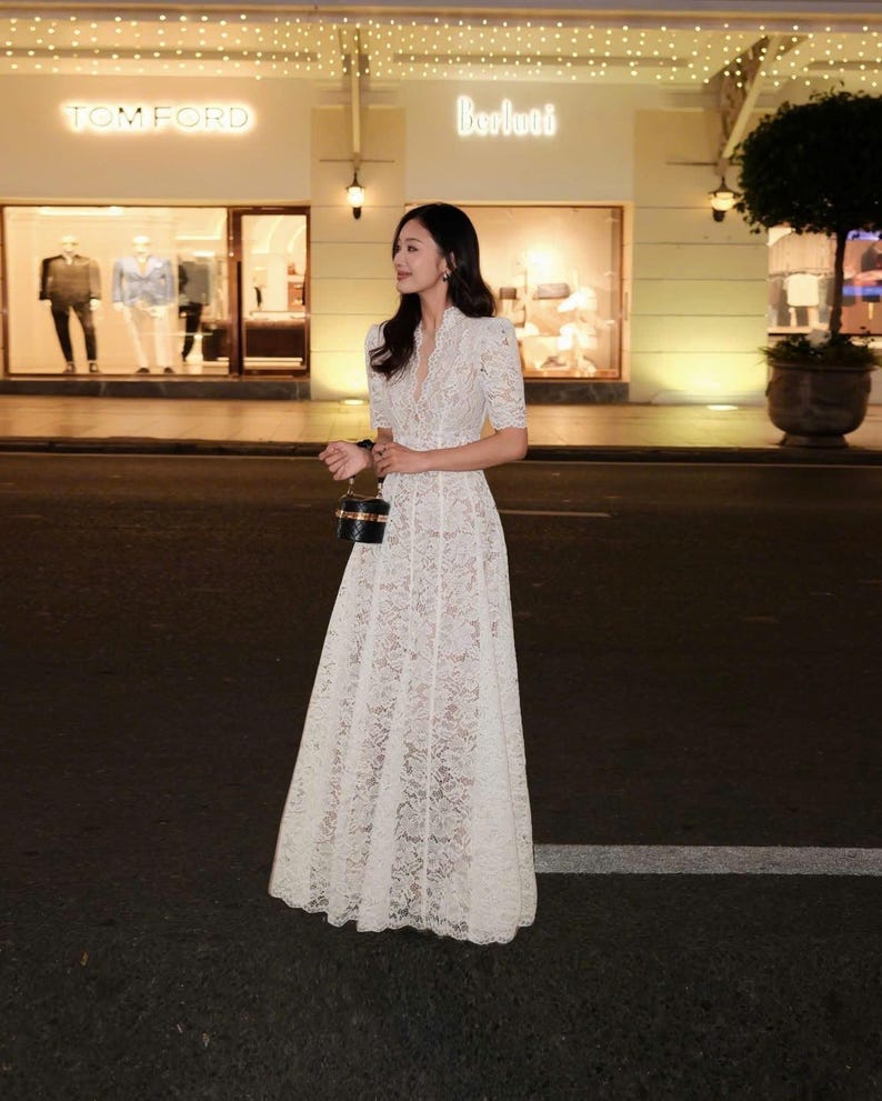 May include: A woman in a white lace dress with short sleeves and a V-neck. The dress has a long, flowing skirt. She is carrying a small, black and gold handbag. The background shows a shopfront with the Tom Ford and Berluti logos.