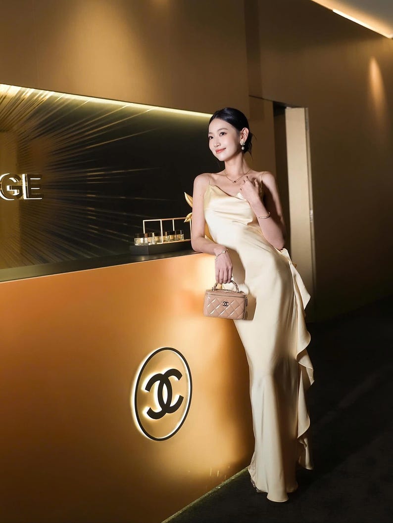 May include: A floor-length, pale yellow satin slip dress with thin spaghetti straps and a ruffled side detail, paired with a small, beige, quilted leather vanity bag featuring a gold-tone interlocking CC logo and a top handle.