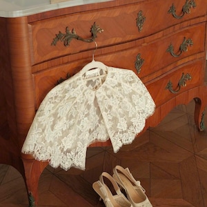 Op de afbeelding: Een cr&egrave;mekleurige kanten capelet met een bloemmotief en geschulpte randen hangend aan een witte hanger, gecombineerd met een bijpassend paar cr&egrave;mekleurige kanten slingback-hakken met spitse neuzen en dunne hakken.