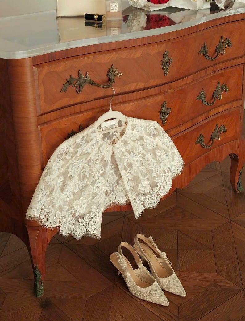 Op de afbeelding: Een cr&egrave;mekleurige kanten capelet met een bloemmotief en geschulpte randen hangend aan een witte hanger, gecombineerd met een bijpassend paar cr&egrave;mekleurige kanten slingback-hakken met spitse neuzen en dunne hakken.