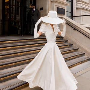 May include: A white midi dress with short puff sleeves, a V-neckline, and a full skirt. The outfit includes a wide-brimmed white hat with a ribbon band, white elbow-length gloves, and white closed-toe heels, creating a coordinated monochromatic look.