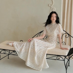 May include: A floor-length cream-colored lace dress featuring a high neckline, short flutter sleeves, and a fitted bodice that transitions into a flowing skirt. The intricate floral lace pattern covers the entire garment, creating a textured look.