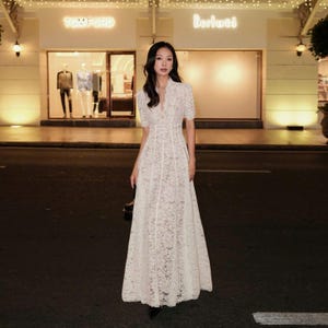 May include: A white lace maxi dress with short sleeves and a V-neck. The dress has a fitted bodice and a flowing skirt. The background includes a storefront with the name "TOM FORD" in illuminated letters.