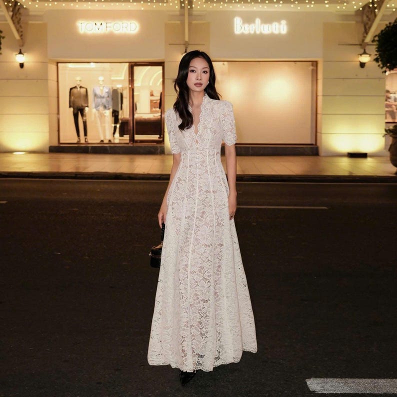 May include: A white lace maxi dress with short sleeves and a V-neck. The dress has a fitted bodice and a flowing skirt. The background includes a storefront with the name "TOM FORD" in illuminated letters.