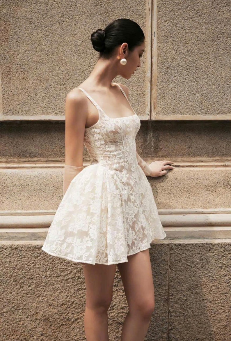 Op de afbeelding: Een korte, mouwloze jurk van off-white kant met een aansluitend lijfje met een vierkante halslijn en een uitlopende rok. Het kledingstuk heeft een gestructureerd silhouet met delicate bloemmotieven in de kanten stof en dunne schouderbandjes.