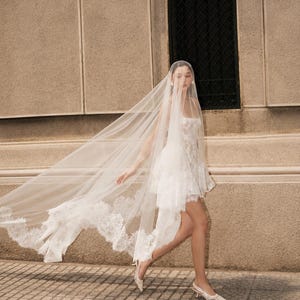 Op de afbeelding: Een witte korte jurk met een vierkante halslijn en kanten details, gecombineerd met een lange, doorschijnende, vloerlange sluier met ingewikkelde kanten randen. Het geheel wordt gecompleteerd door lichtgekleurde slingback-pumps met puntige neus.