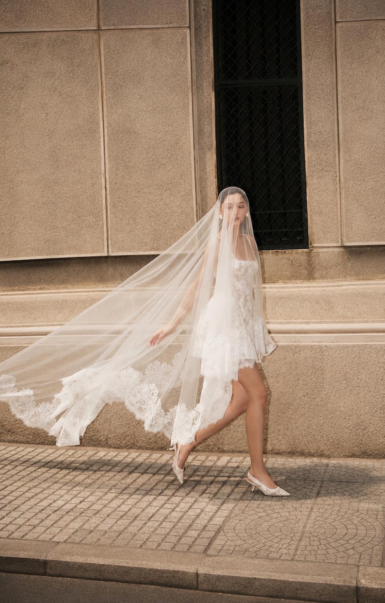 Op de afbeelding: Een witte korte jurk met een vierkante halslijn en kanten details, gecombineerd met een lange, doorschijnende, vloerlange sluier met ingewikkelde kanten randen. Het geheel wordt gecompleteerd door lichtgekleurde slingback-pumps met puntige neus.