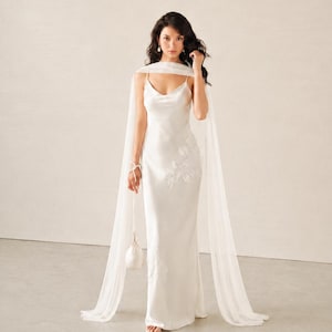May include: A floor-length white satin slip dress with a cowl neckline and thin spaghetti straps. The dress features delicate white floral lace appliqu&eacute;s on the side and a matching sheer white chiffon cape draped over the shoulders.