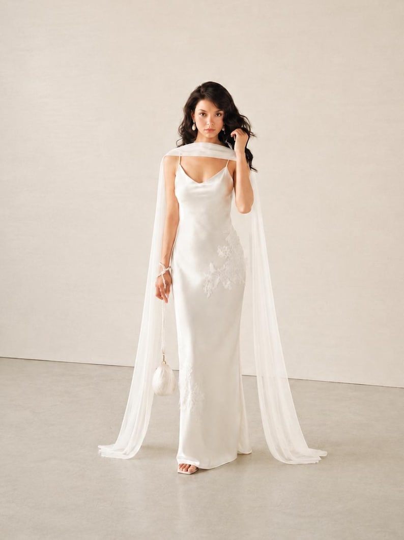 May include: A floor-length white satin slip dress with a cowl neckline and thin spaghetti straps. The dress features delicate white floral lace appliqu&eacute;s on the side and a matching sheer white chiffon cape draped over the shoulders.