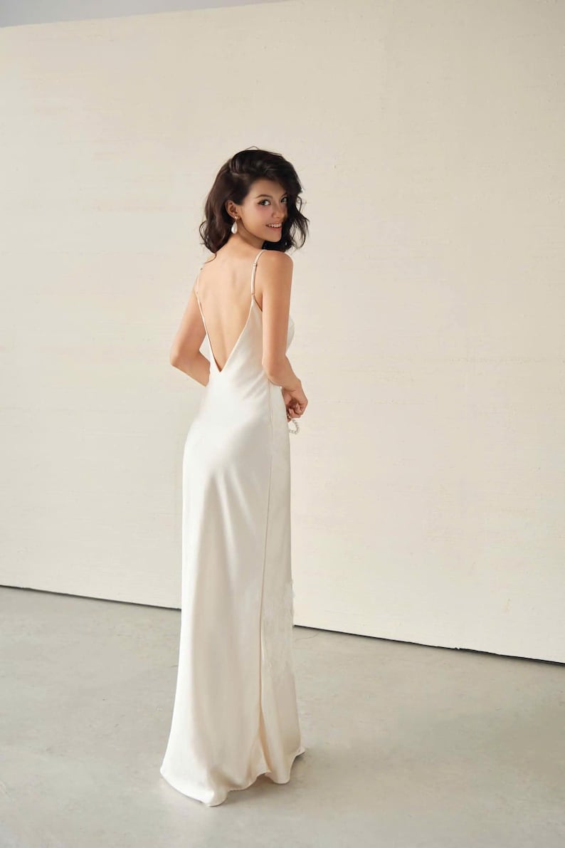 May include: A floor-length, cream-coloured satin slip dress featuring a deep, open back design with thin spaghetti straps. The garment has a smooth, flowing silhouette that extends to the floor, crafted from a lustrous, light-reflecting fabric.
