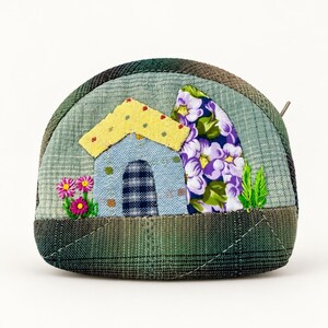 May include: A semi-circular fabric coin purse featuring a patchwork design of a small house with a yellow roof, a blue door, and purple floral accents. The background is a light green plaid fabric, while the bottom section is a dark green plaid.