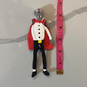May include: A handmade vampire figurine, approximately 3 inches tall, with a red cape, white shirt, black pants, and a yellow button. The vampire has gray skin, red eyes, and a white bow tie. The figurine is standing upright.