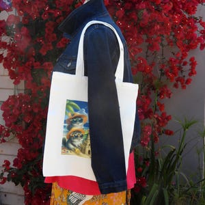 May include: White tote bag with a colorful graphic of three cats wearing sunglasses and hats on a beach. The bag is carried by a person wearing a denim jacket, red top, and a yellow skirt with a floral pattern.