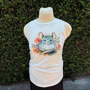 May include: White tank top featuring a watercolor illustration of a chinchilla surrounded by peach-colored roses and greenery. The sleeveless top has a scoop neck and a slightly curved hemline. The chinchilla has gray fur and large ears.