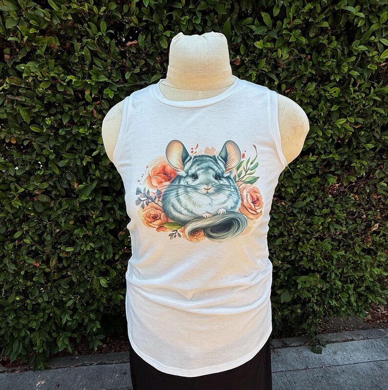 May include: White tank top featuring a watercolor illustration of a chinchilla surrounded by peach-colored roses and greenery. The sleeveless top has a scoop neck and a slightly curved hemline. The chinchilla has gray fur and large ears.