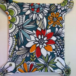 Puede incluir: Una hamaca de tela cuadrada con un vibrante estampado floral en tonos azul verdoso, blanco, naranja, rojo y verde lima. Las esquinas son redondeadas e incluyen ojales metálicos con clips plateados para colgarla de forma segura.