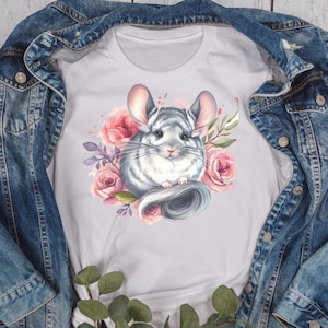 May include: White t-shirt featuring a watercolor illustration of a chinchilla surrounded by pink roses and purple leaves. The chinchilla is gray and white with large ears and a fluffy tail. The shirt is displayed on a denim jacket.