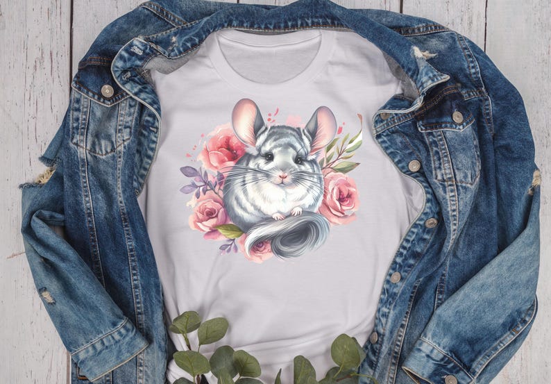 May include: White t-shirt featuring a watercolor illustration of a chinchilla surrounded by pink roses and purple leaves. The chinchilla is gray and white with large ears and a fluffy tail. The shirt is displayed on a denim jacket.