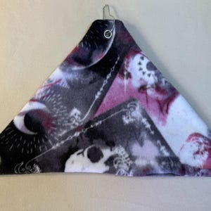Puede incluir: Una hamaca triangular para mascotas hecha de tela polar suave con un patrón oscuro de calaveras, lunas crecientes y adornos en tonos negro, blanco y magenta. Las esquinas incluyen ojales metálicos con ganchos de metal plateado unidos.