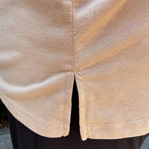 May include: Close-up of a beige-colored shirt with a split hem. The shirt's hemline is visible, with a double-stitched seam. The fabric appears to be a soft, knit material. The shirt is paired with black pants.