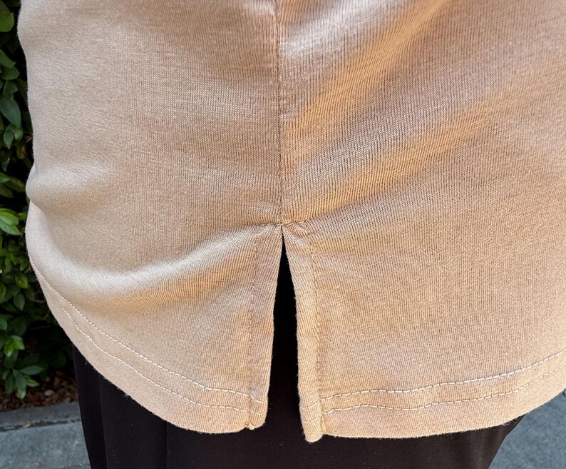 May include: Close-up of a beige-colored shirt with a split hem. The shirt's hemline is visible, with a double-stitched seam. The fabric appears to be a soft, knit material. The shirt is paired with black pants.
