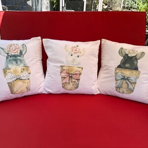 May include: Three white throw pillows with a red couch in the background. Each pillow features a cartoon illustration of a chinchilla wearing a flower crown and sitting in a pot with a bow. The chinchillas are gray, white, and black.