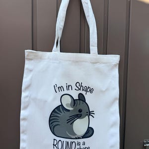 May include: White tote bag with a cartoon chinchilla illustration. The chinchilla is gray and white with the text "I'm in Shape" and "ROUND is a shape" printed on the front. The bag has two white handles.