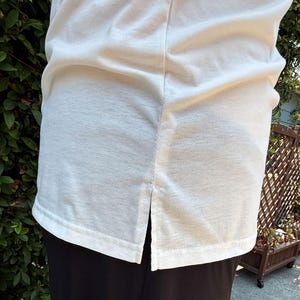 May include: Cream-colored, short-sleeved shirt with a split hem at the bottom. The shirt is made of a soft, textured fabric. The bottom of the shirt is visible, along with the top of the black pants.
