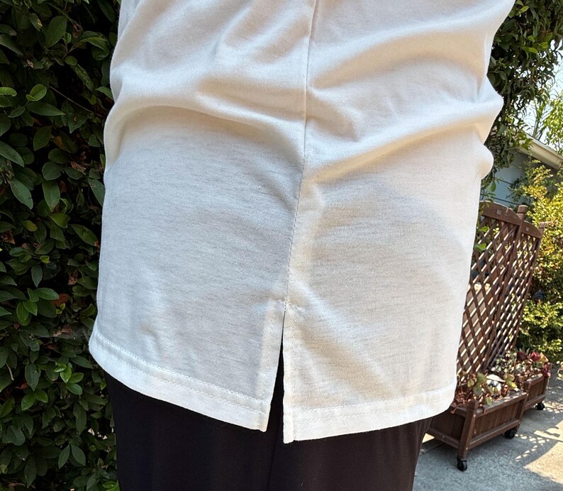 May include: Cream-colored, short-sleeved shirt with a split hem at the bottom. The shirt is made of a soft, textured fabric. The bottom of the shirt is visible, along with the top of the black pants.