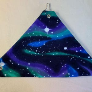 Puede incluir: Una hamaca triangular de tela con un estampado espacial oscuro que muestra nubes de nebulosa en tonos morado y turquesa con estrellas blancas. Las esquinas están reforzadas con ojales metálicos plateados, cada uno con un gancho metálico.