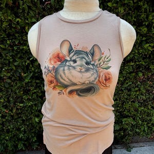 May include: A tan sleeveless shirt featuring a detailed illustration of a chinchilla surrounded by peach-colored roses and green foliage. The chinchilla has gray and white fur and large ears. The shirt is displayed on a mannequin.