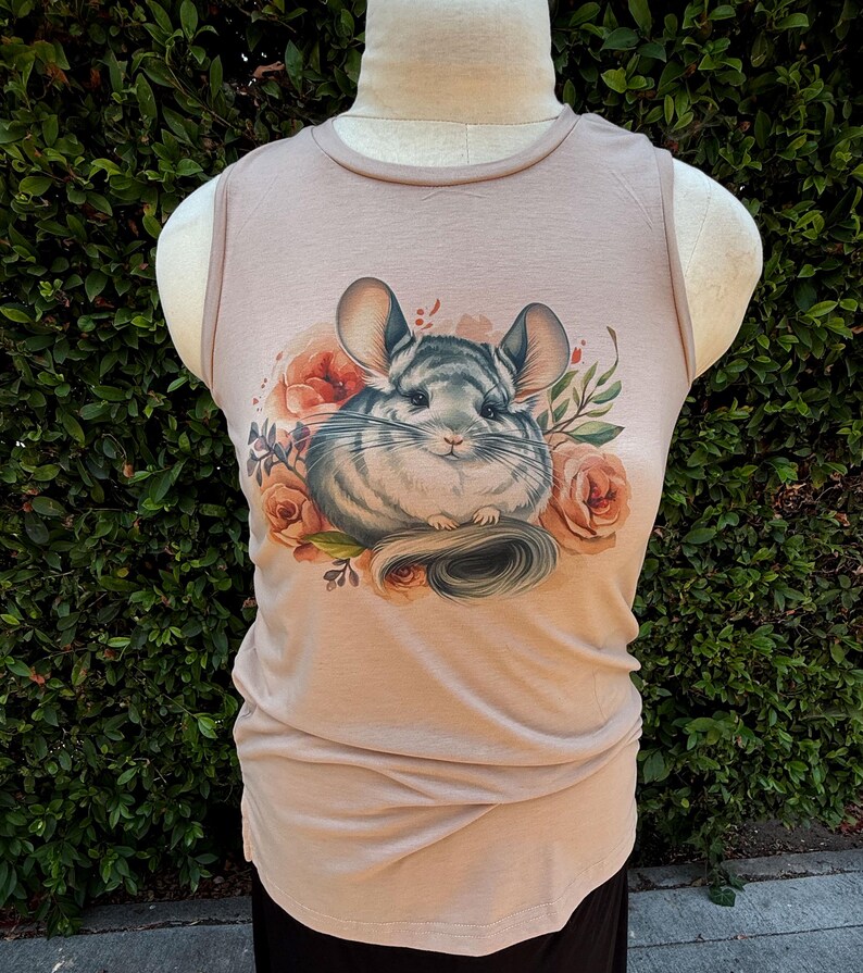 May include: A tan sleeveless shirt featuring a detailed illustration of a chinchilla surrounded by peach-colored roses and green foliage. The chinchilla has gray and white fur and large ears. The shirt is displayed on a mannequin.