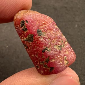 Puede incluir: Una piedra preciosa de rubí en bruto y sin pulir con un color rojo intenso y una superficie irregular y texturizada. La piedra presenta prominentes inclusiones minerales de color negro oscuro dispersas por su superficie, creando un aspecto natural y rugoso.