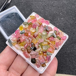 Puede incluir: Una colección de pequeñas piedras preciosas de turmalina en bruto y multicolor en tonos rosa, verde, amarillo y marrón. Estos fragmentos minerales translúcidos de forma irregular están contenidos en una caja expositora cuadrada blanca.