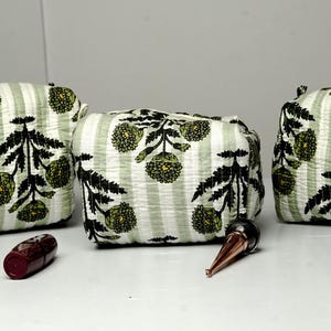 Puede incluir: Tres neceseres con un estampado de rayas verdes y blancas y un diseño floral negro. Las bolsas van acompañadas de un frasco de esmalte de uñas, una barra de labios y un brillo de labios.