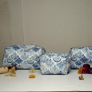 Puede incluir: Tres neceseres a juego con un estampado de conchas marinas en azul y blanco. Varios productos de belleza y accesorios para el cabello están colocados alrededor de los neceseres, incluyendo delineador de ojos, base de maquillaje y pinzas para el pelo. Los neceseres son de diferentes tamaños.