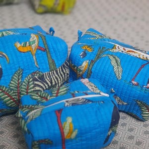 Puede incluir: Tres estuches azules con cremallera con estampado de aves tropicales y cebra. Los estuches son de varios tamaños y tienen una textura acolchada. El diseño incluye tucanes, cebras y follaje tropical sobre un fondo azul.