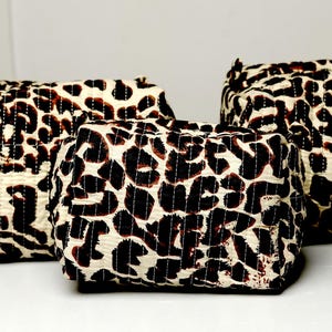 Puede incluir: Tres neceseres con estampado de leopardo de diferentes tamaños. Las bolsas son rectangulares con bordes redondeados y pequeños lazos. El patrón es una mezcla de manchas marrones y negras sobre un fondo crema. Las bolsas están hechas de tela acolchada.
