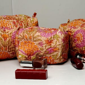 Puede incluir: Juego de tres neceseres con estampado floral de diferentes tamaños. Las bolsas presentan un diseño acolchado con un fondo melocotón y estampados florales naranjas y morados. Artículos de maquillaje, como pintalabios y esmalte de uñas, están al frente.