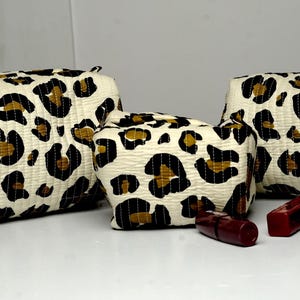 Puede incluir: Tres neceseres con estampado de leopardo de diferentes tamaños, con manchas negras y marrones sobre un fondo blanquecino. Varios artículos de maquillaje, incluyendo barras de labios y un pincel, están delante de los neceseres.